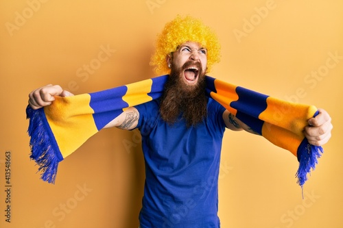 Redhead man with long beard football hooligan cheering game wearing funny wig angry and mad screaming frustrated and furious, shouting with anger. rage and aggressive concept.