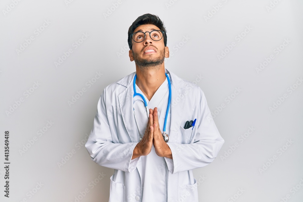 Young handsome man wearing doctor uniform and stethoscope begging and praying with hands together with hope expression on face very emotional and worried. begging.