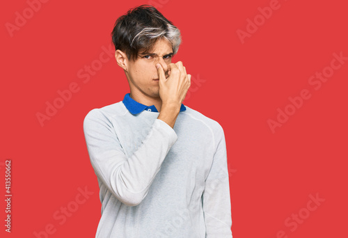 Young hispanic man wearing casual clothes smelling something stinky and disgusting, intolerable smell, holding breath with fingers on nose. bad smell