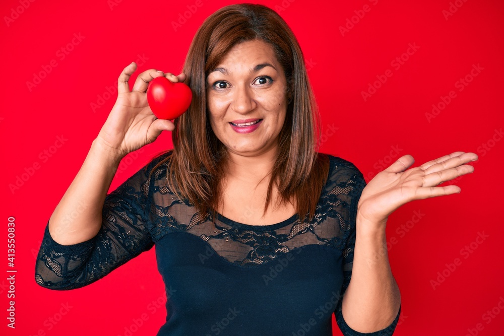 Middle age brunette woman holding heart celebrating achievement with happy smile and winner expression with raised hand Middle age brunette woman holding heart celebrating achievement with happy smile and winner expression with raised hand