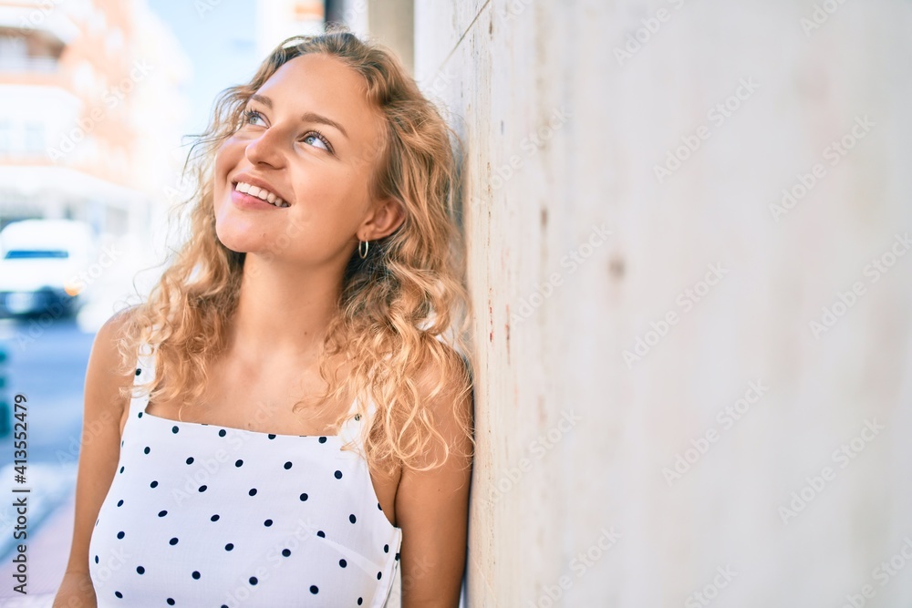 Naklejka premium Young beautiful caucasian woman with blond hair smiling happy outdoors on a summer day leaning on the wall