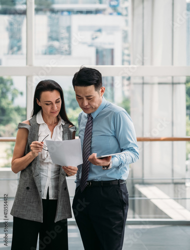 Two business people reading papers together in modern office with big windows.
Two serious business people standing and talking about documents in office lobby