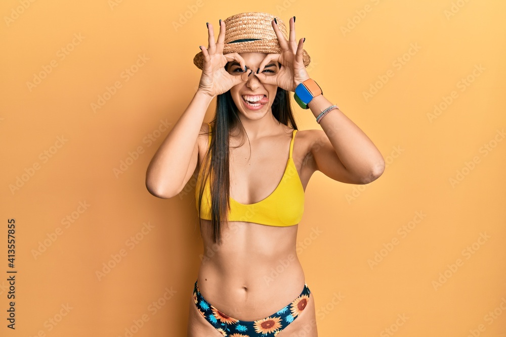 Young hispanic woman wearing bikini and summer hat doing ok gesture like binoculars sticking tongue out, eyes looking through fingers. crazy expression. Young hispanic woman wearing bikini and summer hat doing ok gesture like binoculars sticking tongue out, eyes looking through fingers. crazy expression.