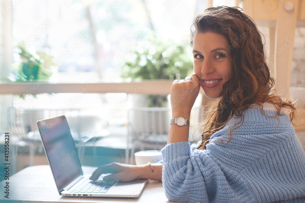Young woman with long hair in blue knitted warm sweater sitting near the window and working on computer Young woman with long hair in blue knitted warm sweater sitting near the window and working on computer