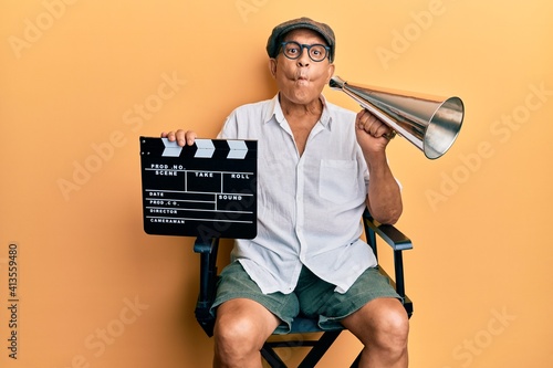 Handsome mature director man holding video film clapboard and louder making fish face with mouth and squinting eyes, crazy and comical. photo