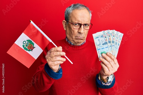 Handsome senior man with grey hair holding peru flag and peruvian sol banknotes skeptic and nervous, frowning upset because of problem. negative person.