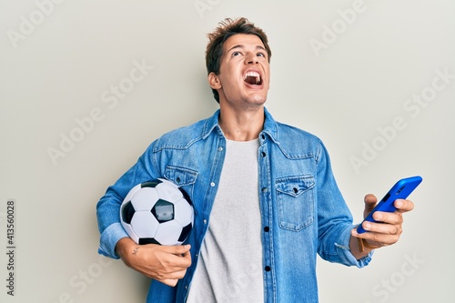 Handsome caucasian man holding football ball looking at smartphone angry and mad screaming frustrated and furious, shouting with anger looking up.