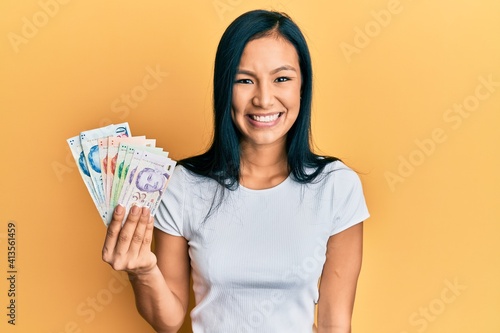 Beautiful hispanic woman holding singapore dollars banknotes looking positive and happy standing and smiling with a confident smile showing teeth