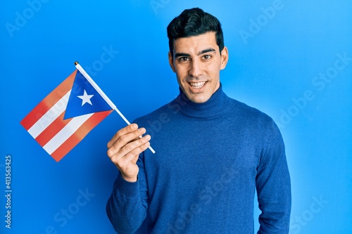 Handsome hispanic man holding puerto rico flag looking positive and happy standing and smiling with a confident smile showing teeth