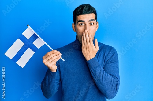 Canvas Print Handsome hispanic man holding finland flag covering mouth with hand, shocked and afraid for mistake