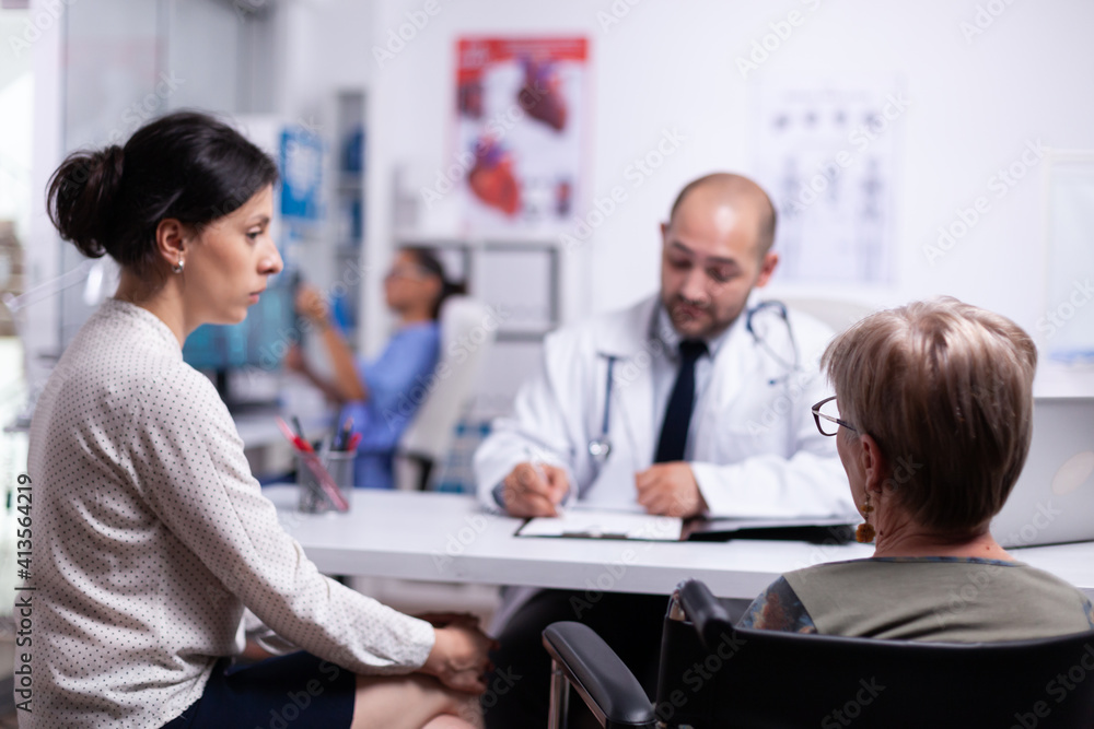 Doughter and disabled mother sitting in wheelchair talking with doctor during consultation. Practitioner asking questions to mature patient filling disease history form, prescribing treatment Doughter and disabled mother sitting in wheelchair talking with doctor during consultation. Practitioner asking questions to mature patient filling disease history form, prescribing treatment