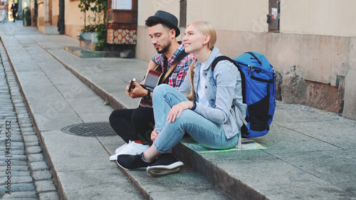 Couple of tourists sitting on sidewalk, playing guitar and having rest. Sightseeing in beautiful european city. © art24pro