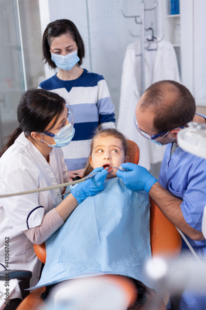 Obraz premium Little girl getting treatment for caries from dentist and assistant wearing dental bib. Mother with her kid in stomatology clinic for teeth examine using modern instruments.