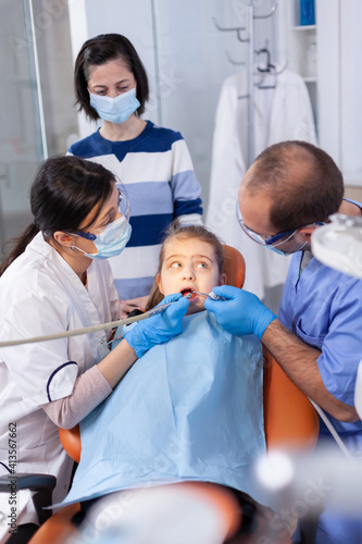 Little girl getting treatment for caries from dentist and assistant wearing dental bib. Mother with her kid in stomatology clinic for teeth examine using modern instruments.