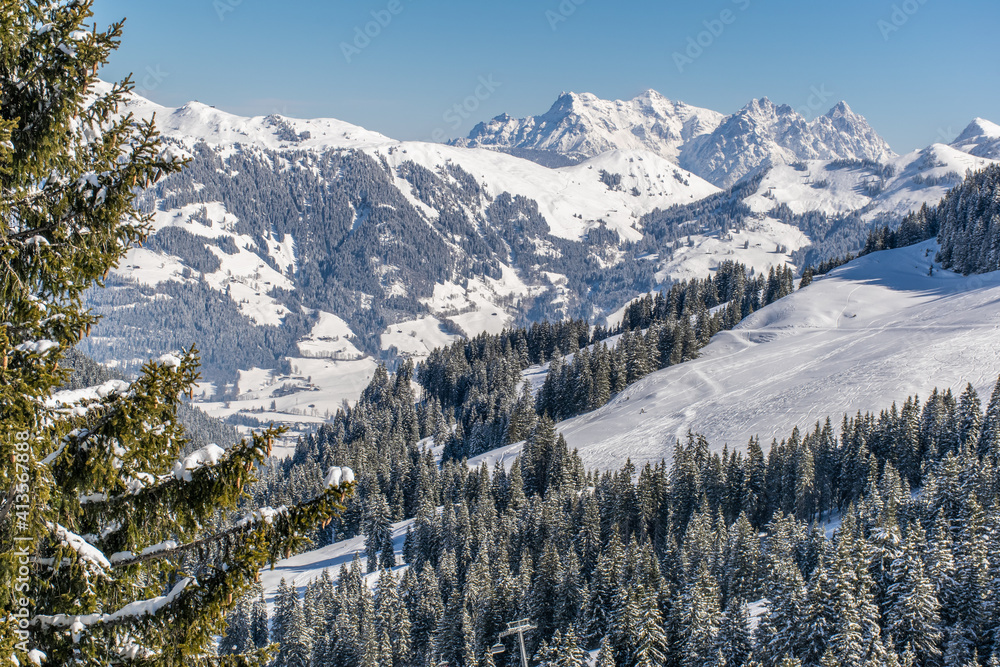The Austrian Alps in winter near Kitzbuhel. Behind the snow-covered fir trees, illuminated by the sun, the magnificent mountain peaks rise against the blue sky.