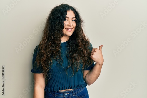 Young brunette woman with curly hair wearing casual clothes smiling with happy face looking and pointing to the side with thumb up.