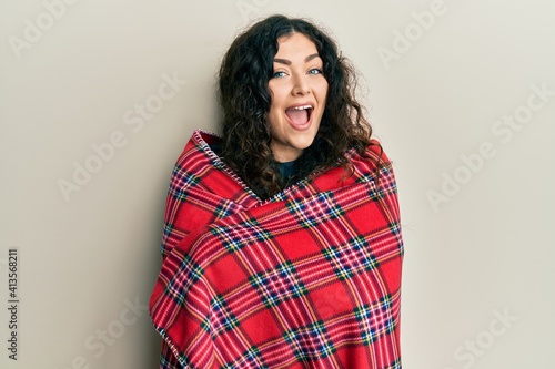 Young brunette woman with curly hair wrapped in a red warm red blanket celebrating crazy and amazed for success with open eyes screaming excited.