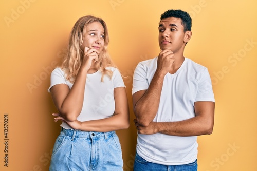 Young interracial couple wearing casual white tshirt with hand on chin thinking about question, pensive expression. smiling with thoughtful face. doubt concept.