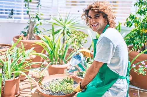 Young hispanic gardener smiling happy caring plants using watering can at terrace