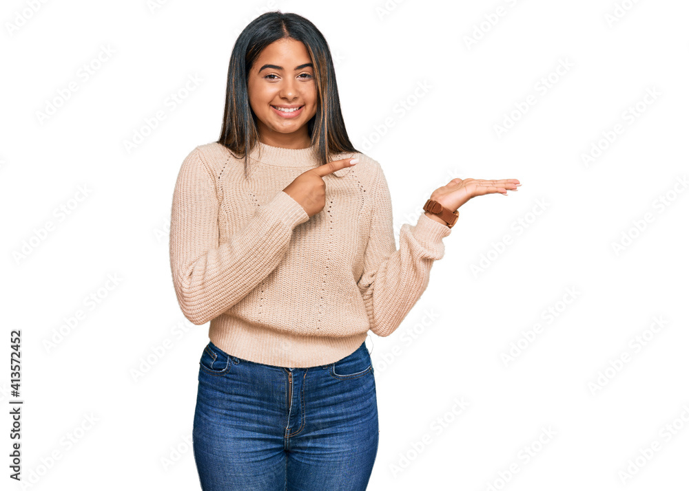 Young latin girl wearing wool winter sweater amazed and smiling to the camera while presenting with hand and pointing with finger. Young latin girl wearing wool winter sweater amazed and smiling to the camera while presenting with hand and pointing with finger.