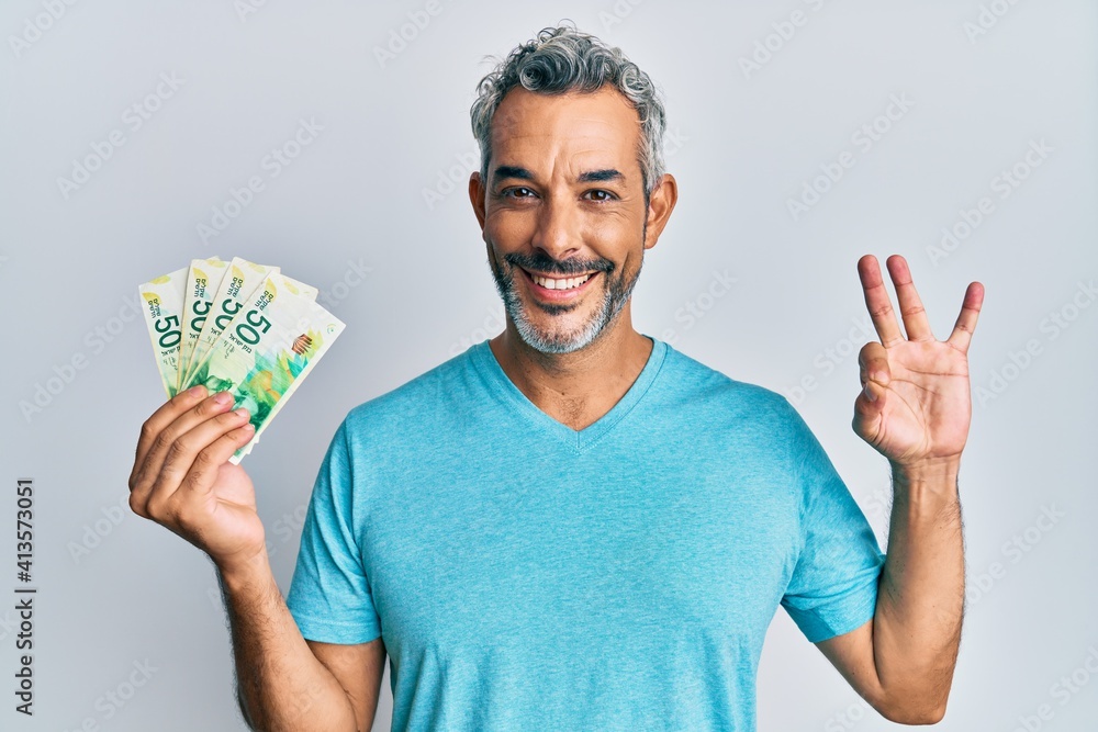 Middle age grey-haired man holding israel shekels banknotes doing ok sign with fingers, smiling friendly gesturing excellent symbol Middle age grey-haired man holding israel shekels banknotes doing ok sign with fingers, smiling friendly gesturing excellent symbol