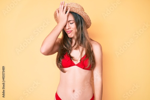 Beautiful young caucasian woman wearing bikini and hat surprised with hand on head for mistake, remember error. forgot, bad memory concept.