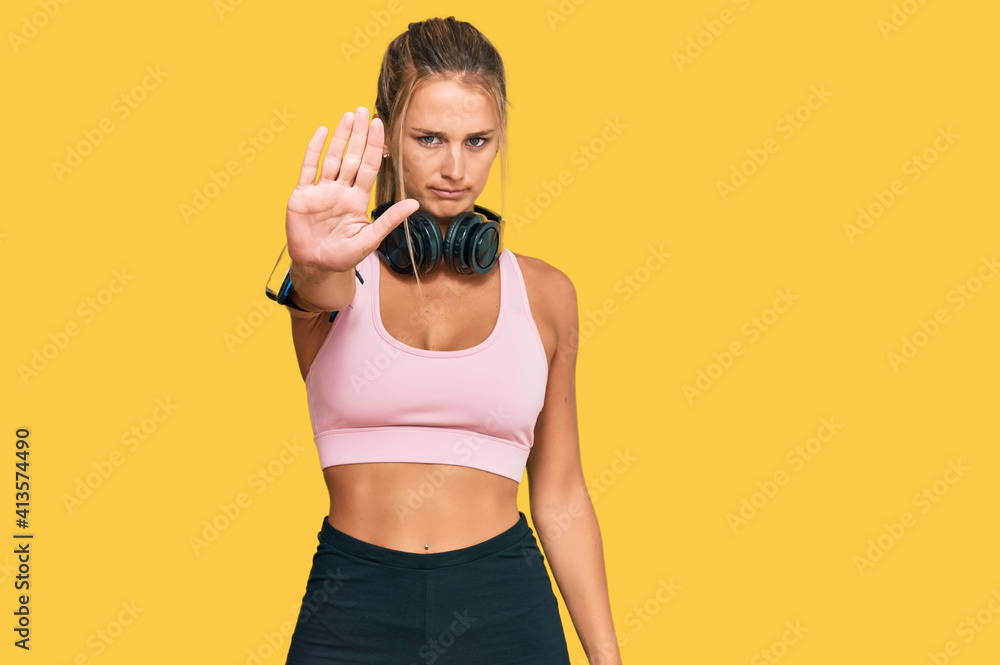 Young blonde woman wearing gym clothes and using headphones doing stop sing with palm of the hand. warning expression with negative and serious gesture on the face. Young blonde woman wearing gym clothes and using headphones doing stop sing with palm of the hand. warning expression with negative and serious gesture on the face.