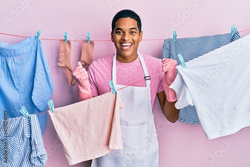 Young handsome hispanic man wearing cleaner apron holding clothes on clothesline celebrating surprised and amazed for success with arms raised and open eyes. winner concept.