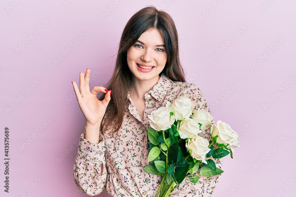 Young beautiful caucasian girl holding bouquet of white flowers doing ok sign with fingers, smiling friendly gesturing excellent symbol Young beautiful caucasian girl holding bouquet of white flowers doing ok sign with fingers, smiling friendly gesturing excellent symbol
