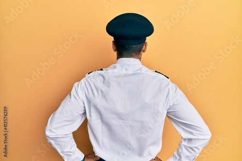 Handsome hispanic man wearing airplane pilot uniform standing backwards looking away with arms on body