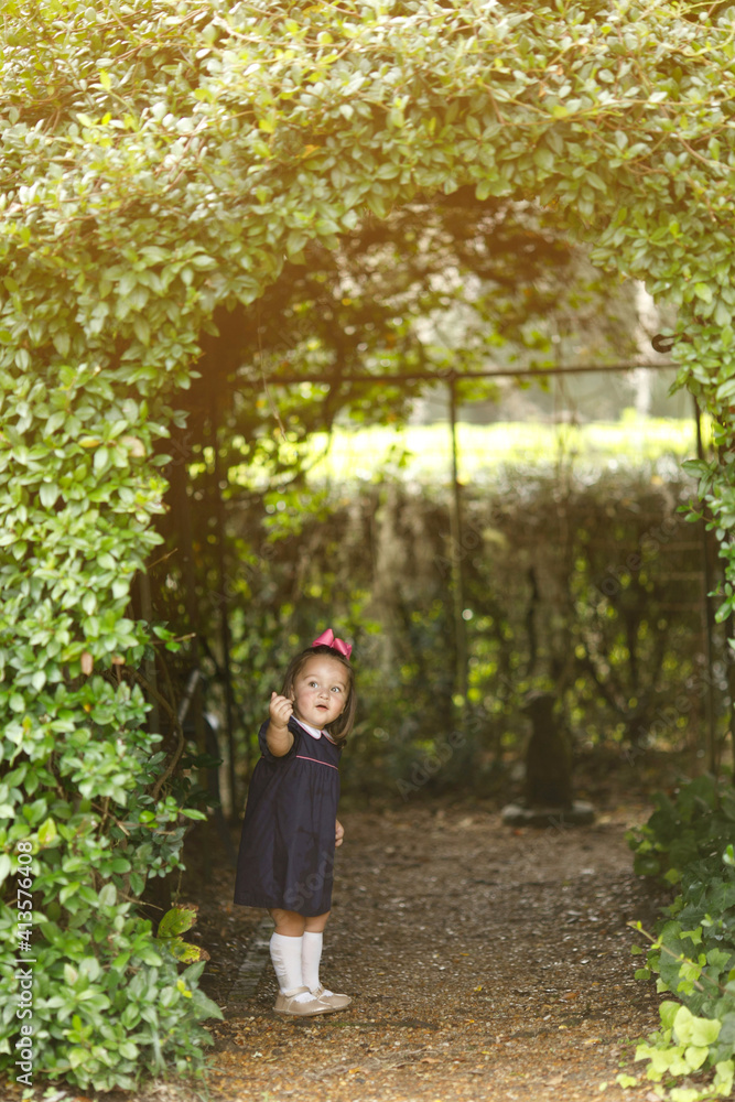 Beautiful preschool toddler aged little girl peeking out from a greenery covered arch