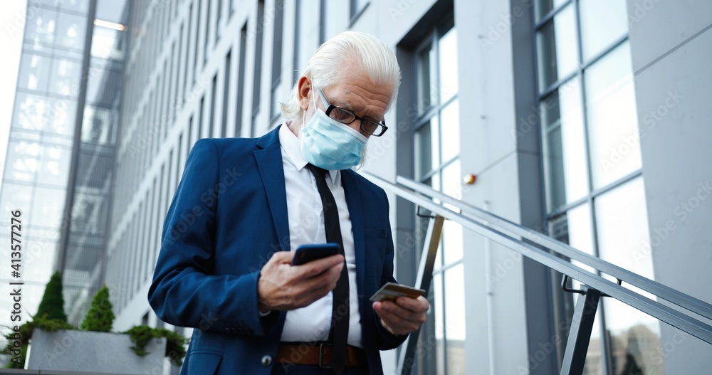 Caucasian old gray-haired man in medical mask using smartphone and credit card to buy online outdoor. Senior businessman pensioner shopping in internet on mobile phone at street. Payment. Coronavirus. Caucasian old gray-haired man in medical mask using smartphone and credit card to buy online outdoor. Senior businessman pensioner shopping in internet on mobile phone at street. Payment. Coronavirus.