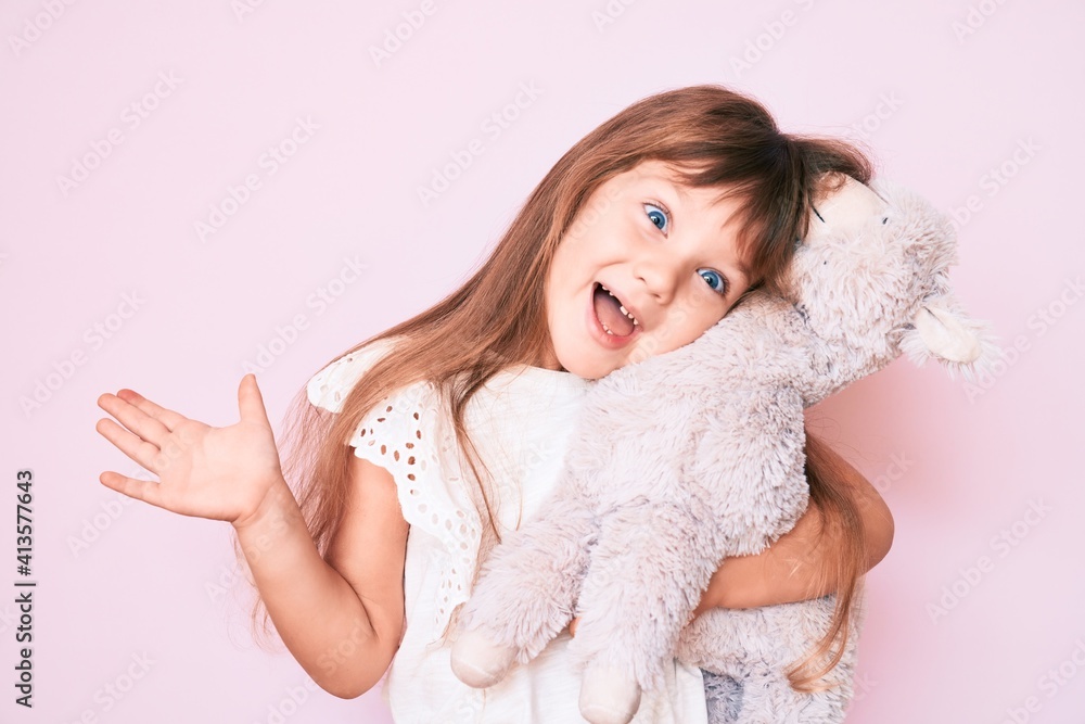 Little caucasian kid girl with long hair holding stuffed animal celebrating achievement with happy smile and winner expression with raised hand Little caucasian kid girl with long hair holding stuffed animal celebrating achievement with happy smile and winner expression with raised hand