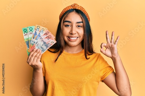 Young latin woman holding australian dollars banknotes doing ok sign with fingers, smiling friendly gesturing excellent symbol