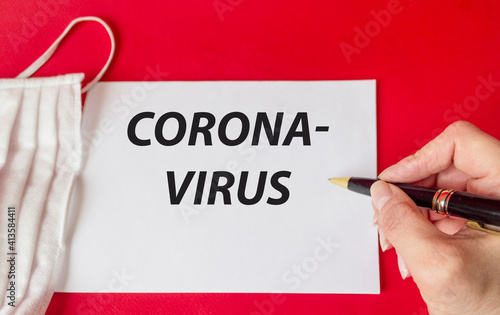 The doctor writes the word Caronovirus on a notebook. A medical mask lies on a red background