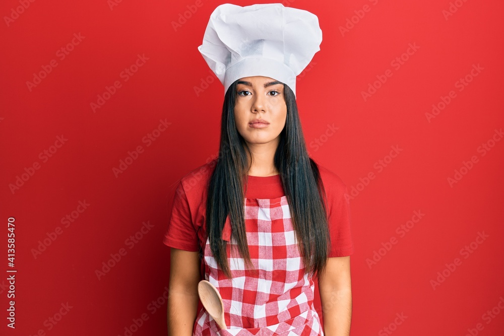 Young beautiful hispanic girl wearing baker uniform and cook hat with serious expression on face. simple and natural looking at the camera. Young beautiful hispanic girl wearing baker uniform and cook hat with serious expression on face. simple and natural looking at the camera.