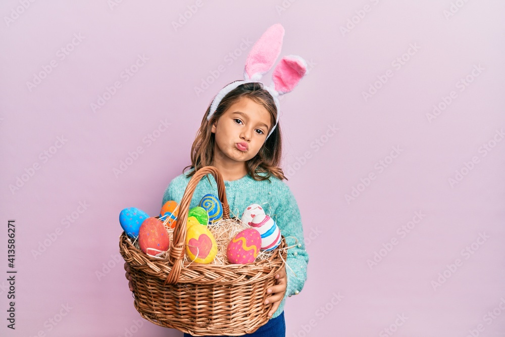 Little beautiful girl wearing cute easter bunny ears holding wicker basket with colored eggs looking at the camera blowing a kiss being lovely and sexy. love expression.