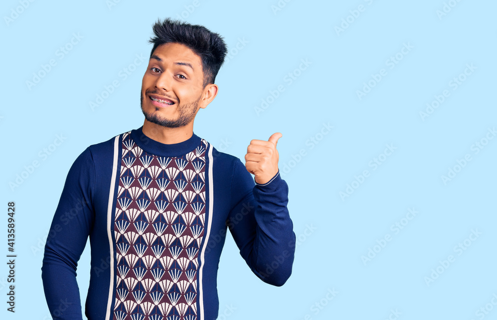 Handsome latin american young man wearing casual winter sweater smiling with happy face looking and pointing to the side with thumb up. Handsome latin american young man wearing casual winter sweater smiling with happy face looking and pointing to the side with thumb up.