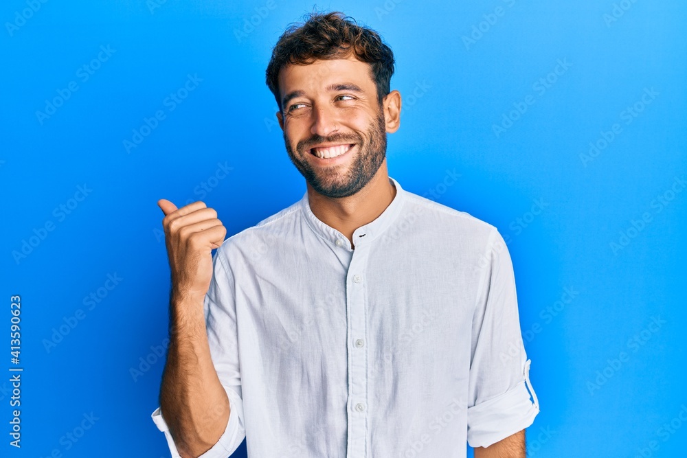 Handsome man with beard wearing elegant shirt pointing thumb up to the side smiling happy with open mouth Handsome man with beard wearing elegant shirt pointing thumb up to the side smiling happy with open mouth