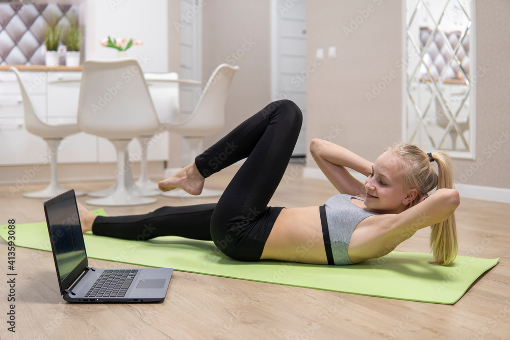 Obraz premium young attractive girl is engaged in fitness at home and looks at the laptop. quarantine fitness concept
