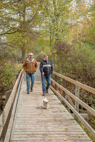 A middle-aged gay couple goes for a walk in the park with their dog in the autumn.