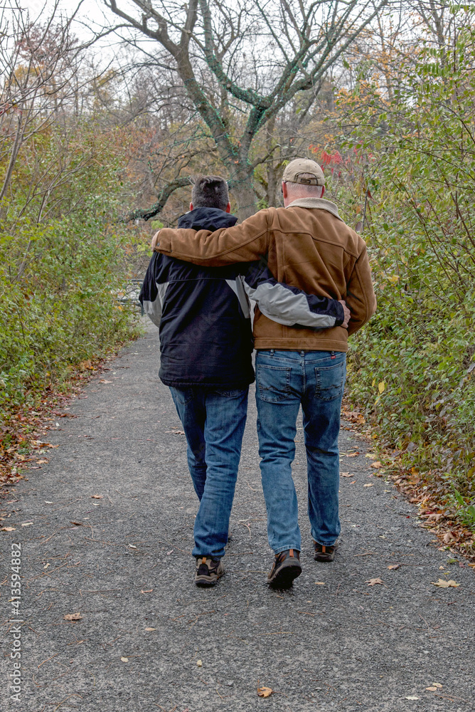 A middle-aged gay couple walking down a path with their arms around each other - backs to camera.
