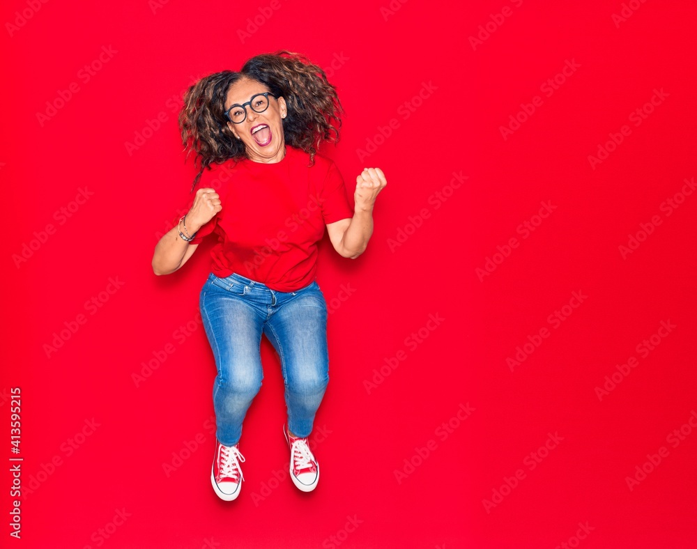Middle age beautiful hispanic woman wearing casual clothes and glasses smiling happy. Jumping with smile on face over isolated red background Middle age beautiful hispanic woman wearing casual clothes and glasses smiling happy. Jumping with smile on face over isolated red background