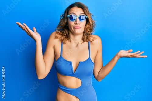 Young caucasian woman wearing swimsuit and sunglasses clueless and confused with open arms, no idea and doubtful face.