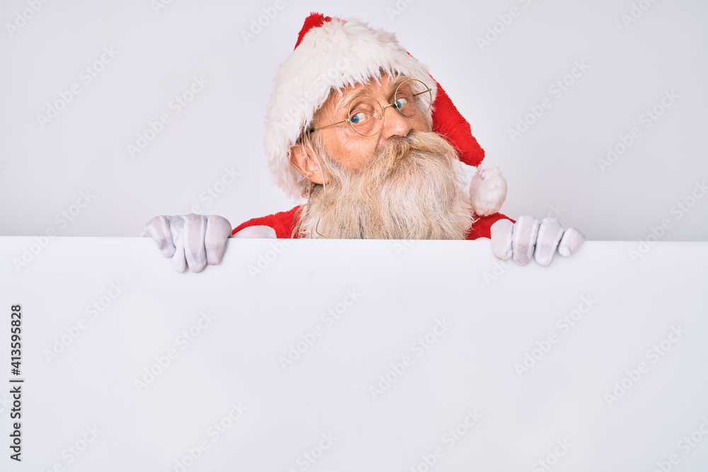 Naklejka premium Old senior man with grey hair and long beard wearing santa claus costume holding banner smiling looking to the side and staring away thinking.