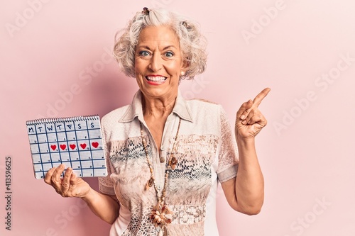 Senior grey-haired woman holding heart calendar smiling happy pointing with hand and finger to the side photo