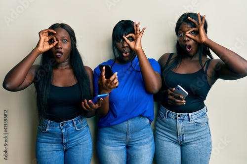 Three young african american friends using smartphone doing ok gesture shocked with surprised face  eye looking through fingers. unbelieving expression.