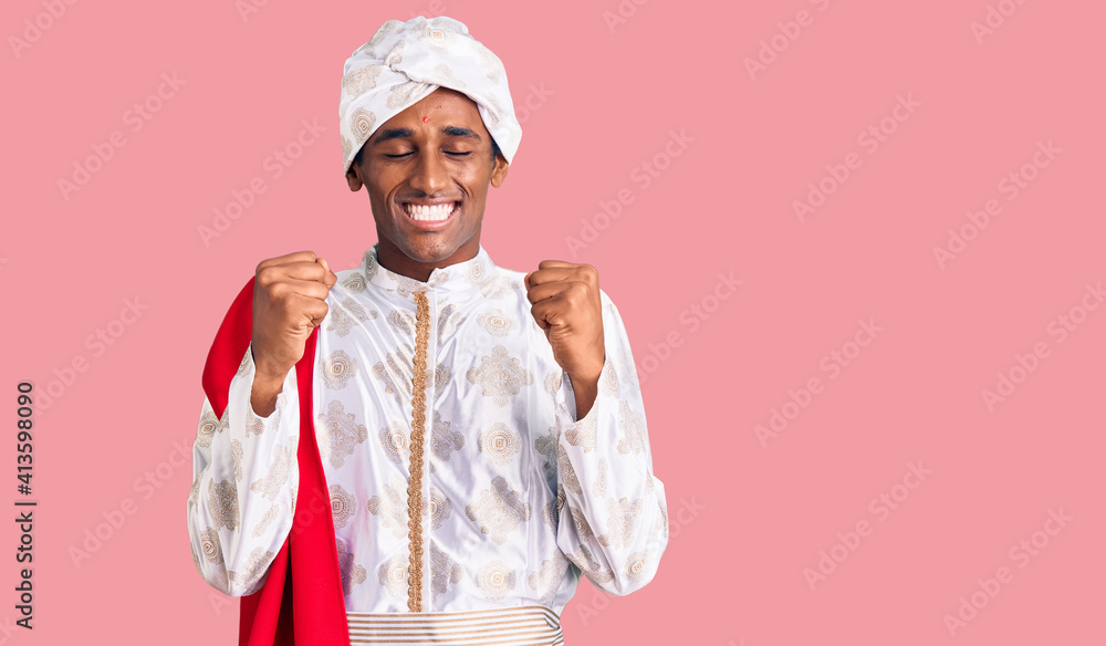 African handsome man wearing tradition sherwani saree clothes excited for success with arms raised and eyes closed celebrating victory smiling. winner concept. African handsome man wearing tradition sherwani saree clothes excited for success with arms raised and eyes closed celebrating victory smiling. winner concept.