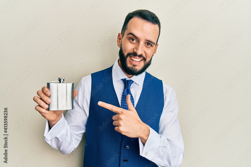 Young man with beard drinking whiskey from flask smiling happy pointing with hand and finger Young man with beard drinking whiskey from flask smiling happy pointing with hand and finger