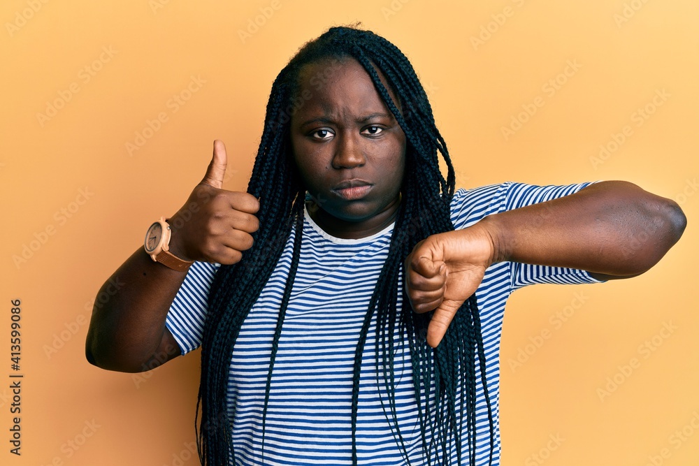 Young black woman with braids doing thumbs down and thumbs up gesture skeptic and nervous, frowning upset because of problem. negative person. Young black woman with braids doing thumbs down and thumbs up gesture skeptic and nervous, frowning upset because of problem. negative person.